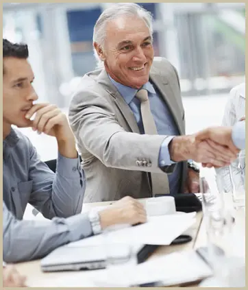 An older man in a suit smiles while shaking hands with another person across a conference table during a meeting. A younger man sits nearby with his hand on his chin.